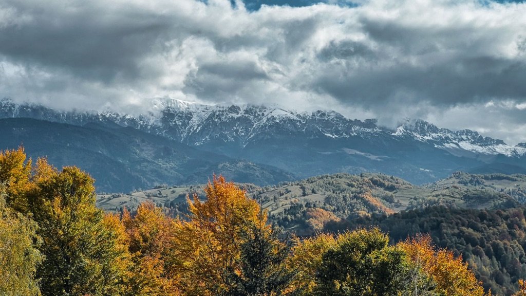 Munți cu zăpadă pe creste, sub nori, deasupra pădurilor colorate de toamnă