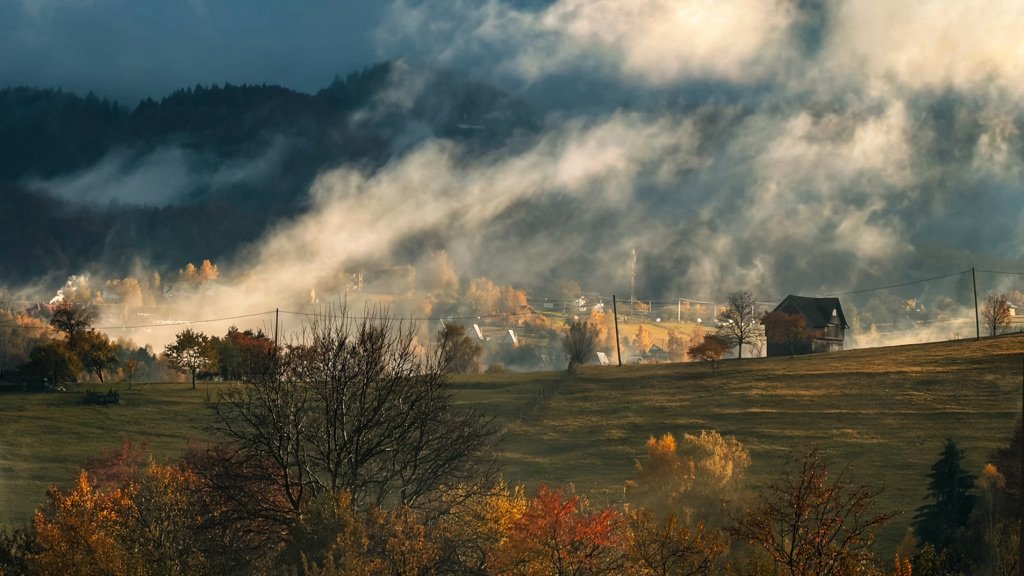 Ceață de dimineață peste un sat de munte, cu copaci în culori de toamnă și pădure de brazi