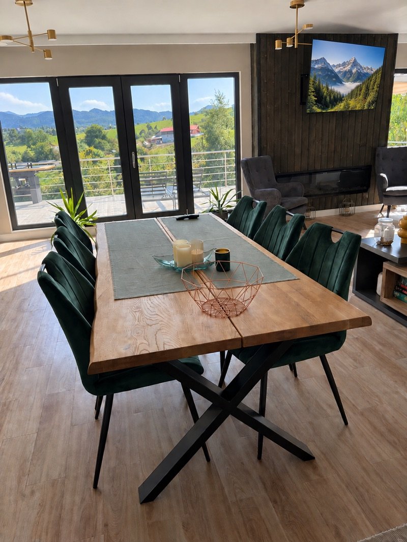 Masa de dining din lemn masiv, cu scaune verzi și ferestre spre dealuri